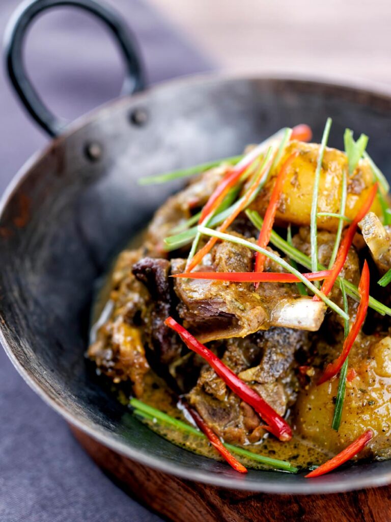 Portrait image of Jamaican curry goat with potatoes served with shredded chilli and green onions served in an iron bowl