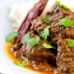 Portrait close up image of an achar gosht lamb or mutton curry served on a white plate with whole chilies and coriander leaves featuring a text overlay