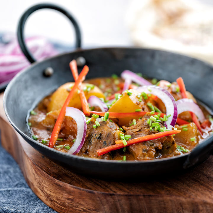 Square image of aloo gosht, an Indian lamb or mutton and potato curry served sliced red onions, snipped chives and sliced red chilli pepper