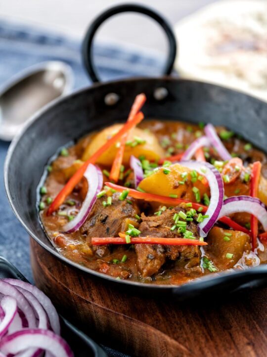 Portrait image of aloo gosht, an Indian lamb or mutton and potato curry served with naan bread and sliced red onions