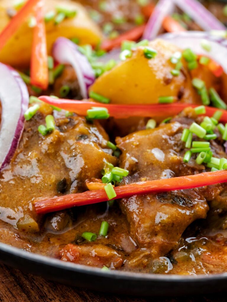 Portrait close image of aloo gosht, an Indian lamb or mutton and potato curry served sliced red onions, snipped chives and sliced red chilli pepper