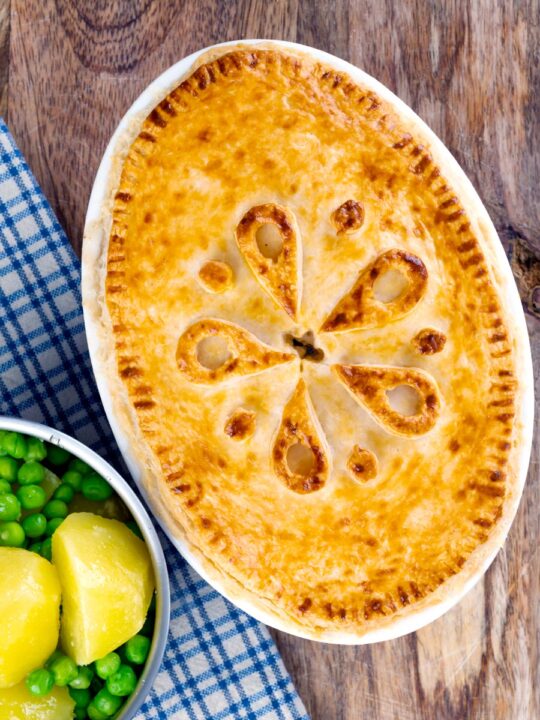 Portrait overhead image of a rabbit pie under a suet crust lid served along side a pan of buttered potatoes and peas.