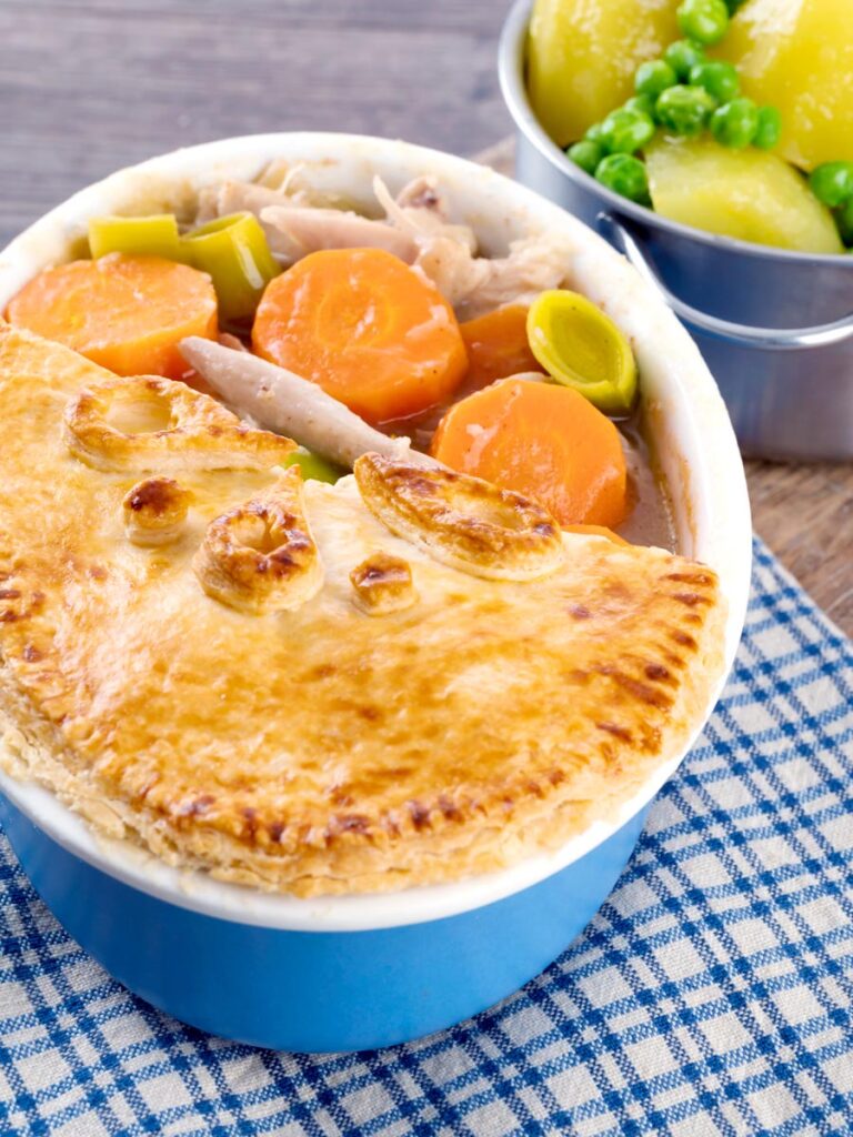 Portrait image of a rabbit pie under a suet crust lid with half removed to show the filling served alongside a pan of buttered potatoes and peas
