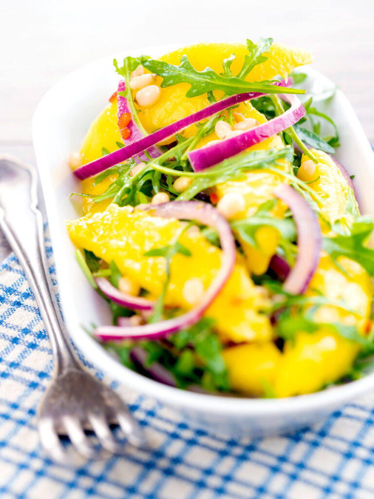 Spicy mango salad with rocket or arugula, shredded red onions and pine nuts served in a white bowl.