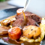 Portrait image of gravy being poured over beer braised beef cheeks with carrot and mushrooms