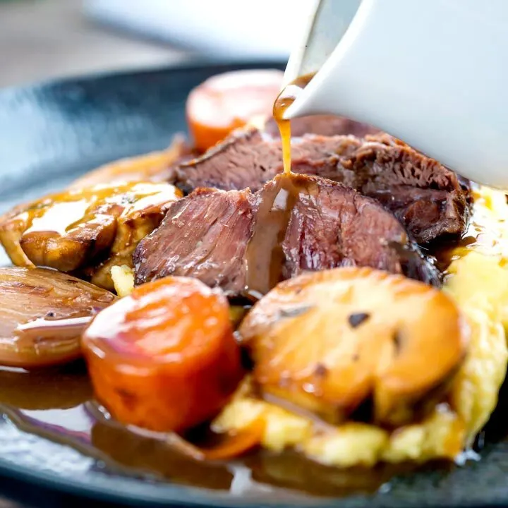 Square image of gravy being poured over beer braised beef cheeks with carrot and mushrooms