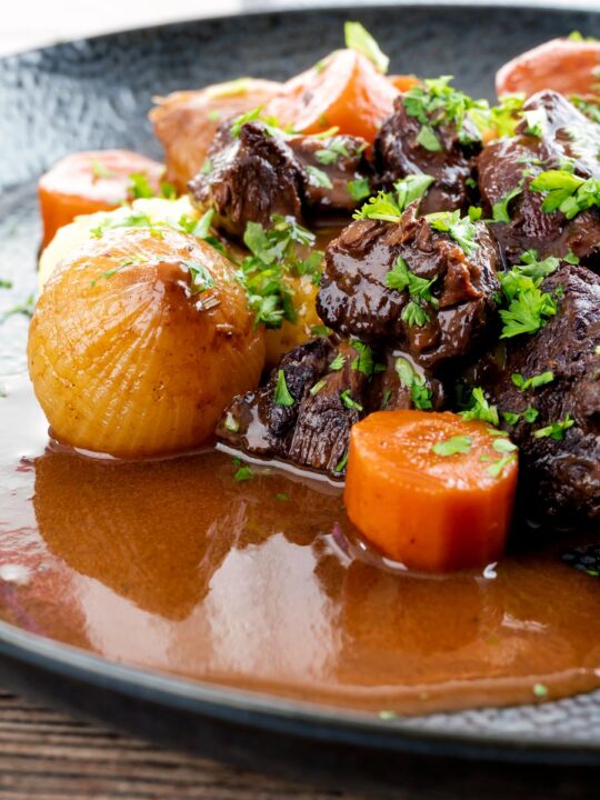 Portrait close up image of a slow cooker beef daube featuring carrots and whole small onions garnished with parsley