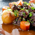 Portrait close up image of a slow cooker beef daube featuring carrots and whole small onions garnished with parsley featuring a title overlay