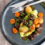 Portrait overhead image of a slow cooker beef daube featuring carrots and whole small onions, served with mashed potato and garnished with parsley
