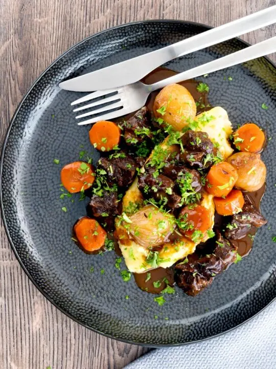 Portrait overhead image of a slow cooker beef daube featuring carrots and whole small onions, served with mashed potato and garnished with parsley