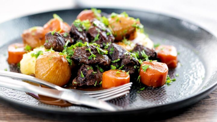 Landscape image of a slow cooker beef daube featuring carrots and whole small onions served with mashed potato and garnished with parsley