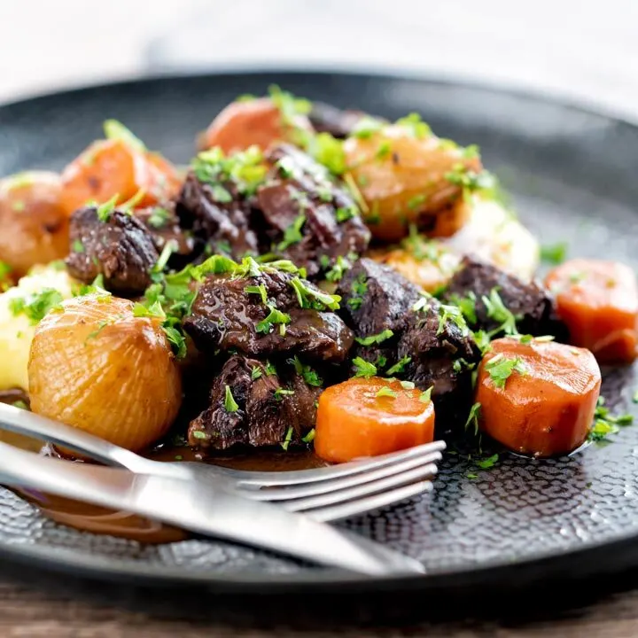 Square image of a slow cooker beef daube featuring carrots and whole small onions served with mashed potato and garnished with parsley