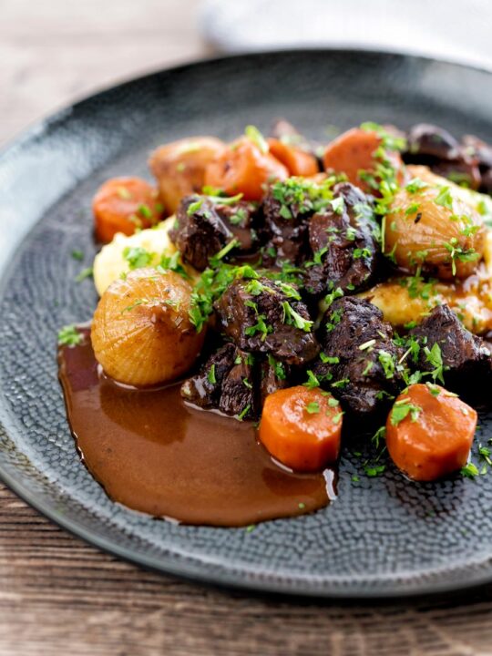 Portrait image of a slow cooker beef daube featuring carrots and whole small onions served with mashed potato and garnished with parsley