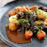 Portrait image of a slow cooker beef daube featuring carrots and whole small onions served with mashed potato and garnished with parsley featuring a title overlay