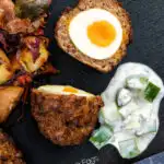 Overhead Nargis kebab or Indian Scotch eggs with one cut open to show jammy yolk served with cucumber raita featuring a title overlay