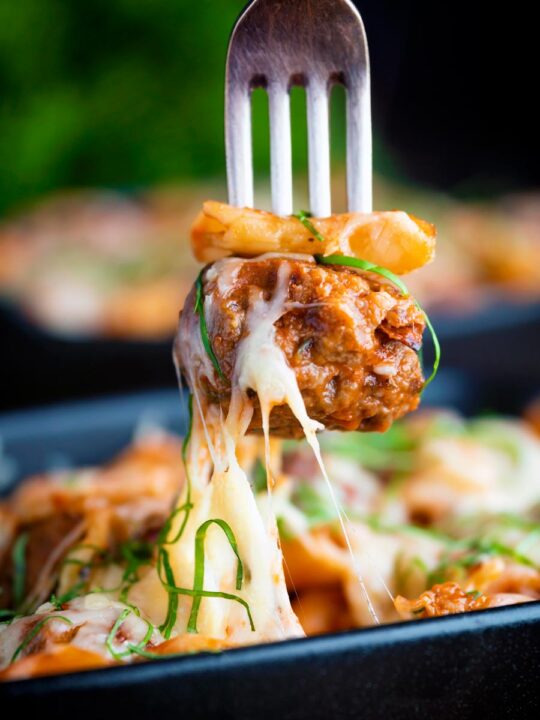 Meatball being removed from a cheesy meatball pasta bake with a fork.