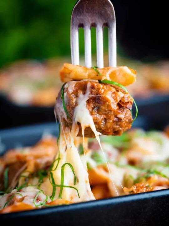 Meatball being removed from a cheesy meatball pasta bake with a fork.