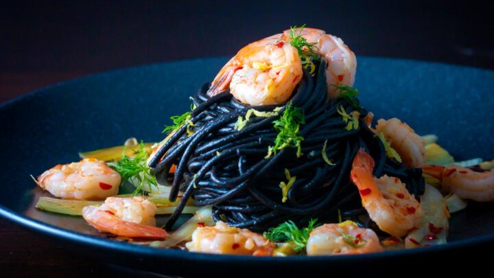 Prawn pasta with squid ink spaghetti fennel, lime zest and chilli served on a black plate.