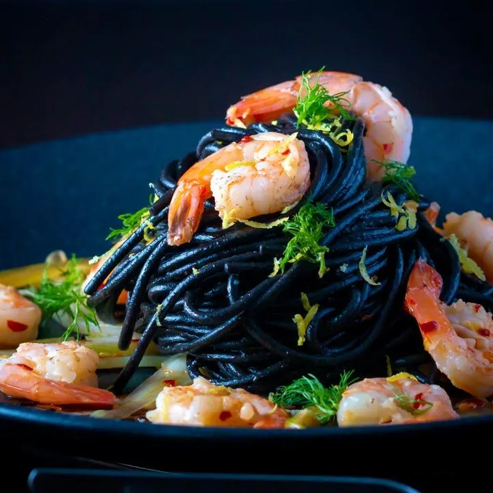 Prawn pasta with squid ink spaghetti fennel, lime zest and chilli served on a black plate.