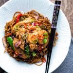 Salt and chilli chicken served with peppers and onion on soy sauce noodles in a white bowl.