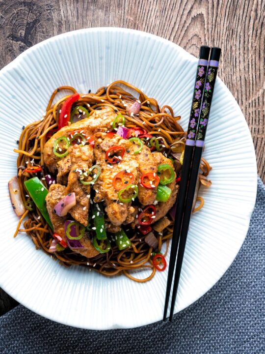 Salt and chilli chicken served with peppers and onion on soy sauce noodles in a white bowl.