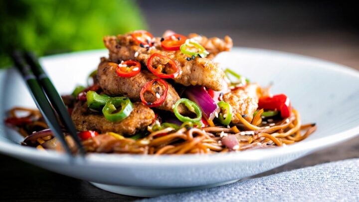 Salt and chilli chicken served with peppers and onion on soy sauce noodles in a white bowl.