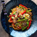 Overhead Szechuan beef stir fry with noodles and bell peppers served in a blue bowl featuring a title overlay