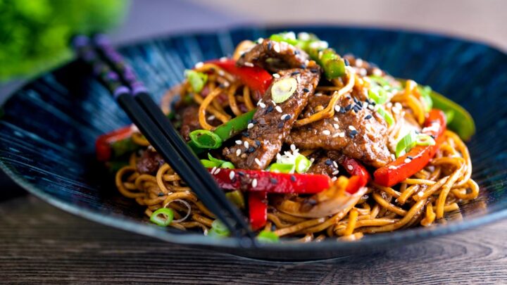 Szechuan beef stir fry with noodles and red and green bell peppers served in a blue bowl.