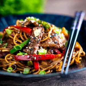 Szechuan beef stir fry with noodles and bell peppers served in a blue bowl with chopsticks.