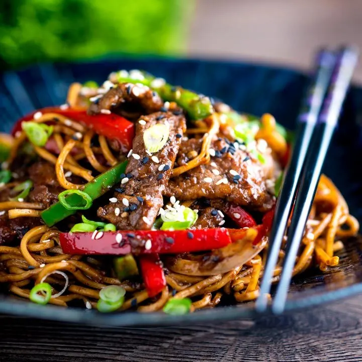 Szechuan beef stir fry with noodles and bell peppers served in a blue bowl with chopsticks.