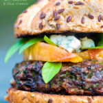 A venison burger served on a bun topped with blue cheese, gin cooked peaches & basil featuring a title overlay.