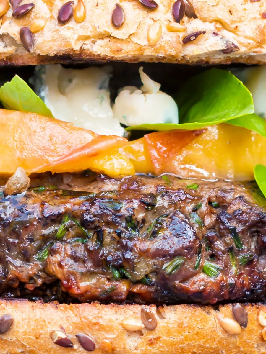 Close up venison burger served on a bun topped with blue cheese, peaches & basil.