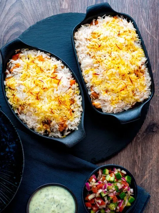 Overhead chicken tikka biryani served in stoneware bowls with kachumber salad and raita.