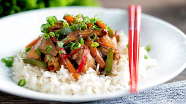 Chicken with black bean sauce serve in a white bowl with rice and spring onions.