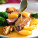 Orange sauce being poured over a rosy pan fried duck breast with crispy skin.