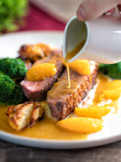 Orange sauce being poured over a rosy pan fried duck breast with crispy skin.