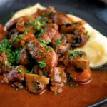 Wild boar stew with mushrooms served with mashed potato on a dark plate.