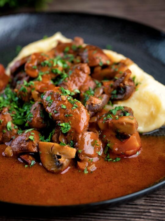 Wild boar stew with mushrooms served with mashed potato on a dark plate.