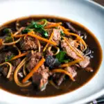 Beef noodle soup featuring udon noodles, spinach & carrot in a dark soy broth featuring a title overlay.