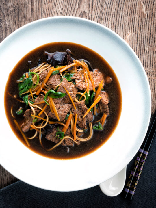 Overhead beef noodle soup featuring udon noodles & spinach in a dark soy broth