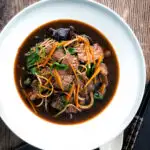 Overhead beef noodle soup featuring udon noodles & spinach in a dark soy broth featuring a title overlay.