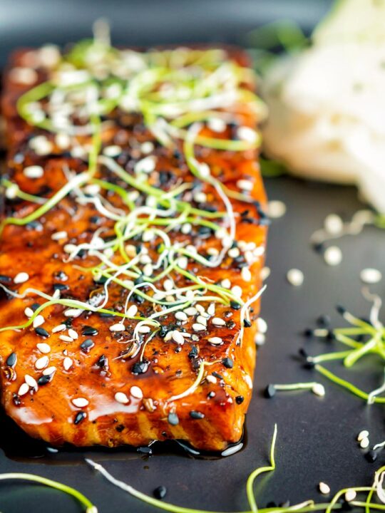 Close up honey soy salmon served on a black plate with pickled daikon and onion sprouts.