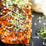 Close up honey soy salmon served on a black plate with pickled daikon and onion sprouts featuring a title overlay.