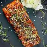 Overhead honey soy salmon served on a black plate with pickled daikon and onion sprouts.