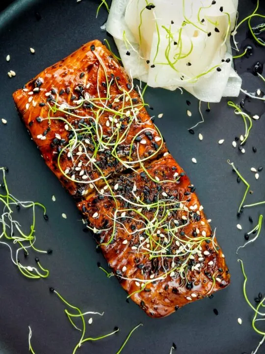 Overhead honey soy salmon served on a black plate with pickled daikon and onion sprouts.