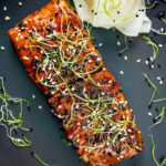 Overhead honey soy salmon served on a black plate with pickled daikon and onion sprouts featuring a title overlay.