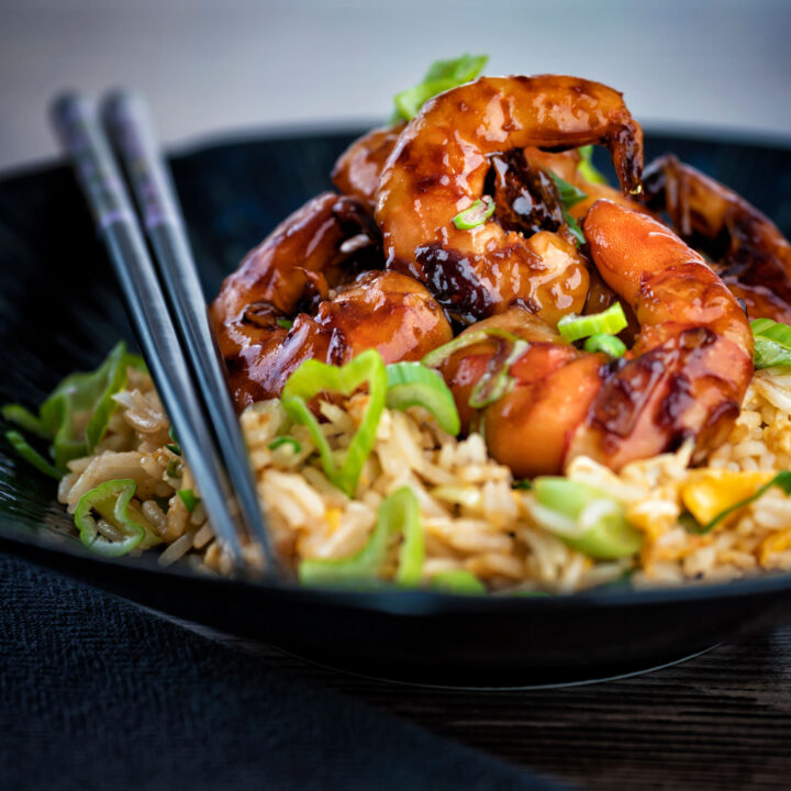 Teriyaki prawns served with egg fried rice and chopsticks on a dark plate.