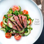 Overhead Thai beef salad with rare beef, tomatoes, mint, chilli and cucumber featuring a title overlay.