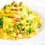 Pasta carbonara with peas and bacon served in a white bowl.