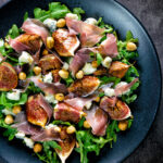 Overhead roasted figs with prosciutto ham, rocket, blue cheese and hazelnuts featuring a title overlay.